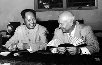 A balding Russian man (Nikita Kruschev) and a younger Chinese man (Mao Zedong) sit and smile, the balding man holding a fan