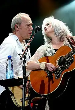Mark Knopfler y Emmylou Harris cantando en un mismo micrófono de pie. Ella toca la guitarra.
