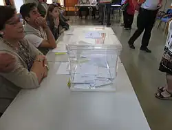 Una mesa electoral con una urna transparente, que contiene varios sobres.
