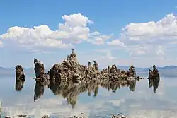 A rock formation rises from the surface of a calm lake. In the distance, the horizon is filled with hills. The sky is blue with clouds The rocks appear grey in colour.