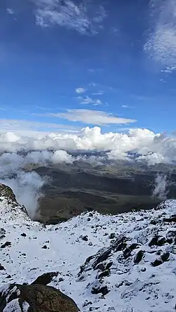 Vista del Nevado del Tolima desde su ladera oriental. En la imagen se observa una amplia cobertura de nieve reciente, típica de las zonas más elevadas del macizo, junto a formaciones rocosas volcánicas y un paisaje de páramo en el horizonte. Este volcán estratovolcánico, ubicado en el Parque Nacional Natural Los Nevados, alcanza una altitud aproximada de 5.215 metros sobre el nivel del mar y conserva uno de los glaciares más pequeños y frágiles de Colombia.