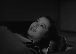 Noriko, vestida con el traje típico, recostada en un futón y cubierta con una manta, mira fijamente al techo y sonríe.