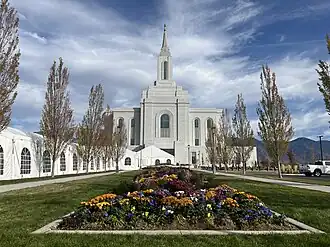 A planted garden in a grounded concrete box is seen in the center, leading to the tall granite parapets of the Orem Utah Temple. On the left, a white tent (from the open house) is seen leading up to the temple entrance.