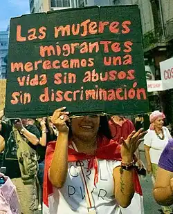 Una mujer joven de entre 20 a 30 años lleva un cartel en una manifestación por el paro internacional de mujeres. El cartel es negro, con letras rojas. En el cartel se lee "Las mujeres migrantes merecemos una vida sin abusos sin discriminación". Alrededor de la mujer hay otras personas caminando y manifestándose. La mujer ocupa el plano central de la foto.