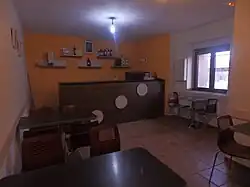 Imagen de una de las salas de la sede de la asociación "Amigos de Olmillos de Muñó" con paredes naranjas donde se puede ver una barra de bar marrón, varias estanterias con botellas, mesas de bar y una ventana