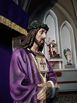 Imagen de 1 metro fabricada en fibra de vidrio de Jesús Nazareno, representando la escena del "Ecce Homo", manos atadas al frente y el pie derecho sobresale ligeramente de la túnica. A su vez porfa un escapulario con la Cruz Trinitaria, una franja azul y superpuesta una franja roja, con el anagrama IHS bordado.