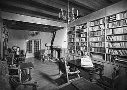 A black and white image of a room with a wood panelled ceiling, with a large fireplace and bookshelves on two sides of the room. At the far end of the room is a glass fronted double door leading away. There are a number of small chairs and tables around the room.