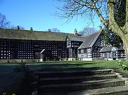 A two-storey black and white timbered building in an L-shape. A wide set of stone steps in the foreground leads onto a grassed area in front of the house.