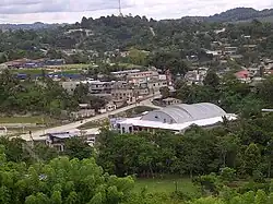 Vista sobre un valle amplio y poco profundo con edificios dispersos intercalados con parches densos de árboles, varios espacios abiertos, cubiertos de hierba. El valle se eleva hacia una cresta baja boscosa. Un mástil de radiodifusión se eleva en una de las colinas.