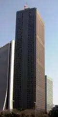 Ground-level view of a brown, rectangular high-rise; the window placement creates several horizontal bands on one side and one vertical stripe on the other