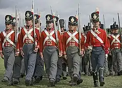 Photo of men dressed in British uniforms of the early 1800s. They wear red coats with white cross belts, gray trousers, and black shakos.