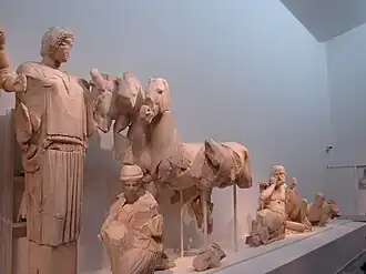 Frontón este, zona derecha. Hipodamía levanta un faldón de su peplo como gesto ritual de la recién casada. Una joven arrodillada se ocupa de los caballos. Un adivino (Clitio, Yamo o Amitaón), muestra en su rostro la angustia al vaticinar el final de la carrera. La figura siguiente es un niño que juega con el dedo pulgar de su pie. Por último, la personificación del río Alfeo (o Kladeos) forma el ángulo del frontón.